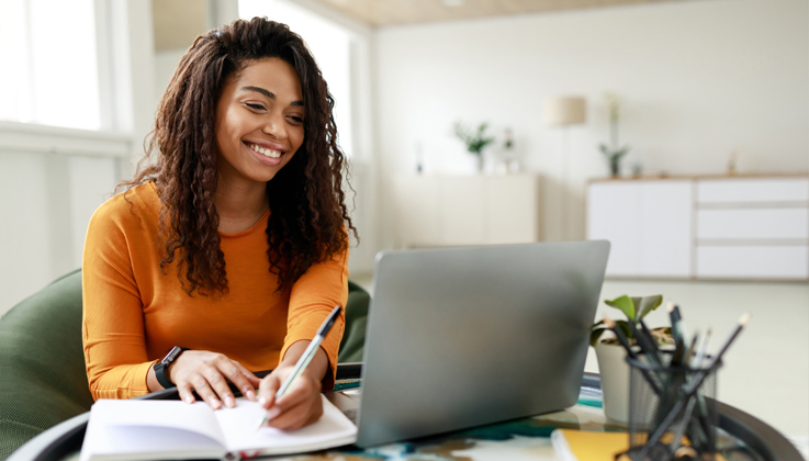 Woman smiling at laptop computer screen while hand-writing notes